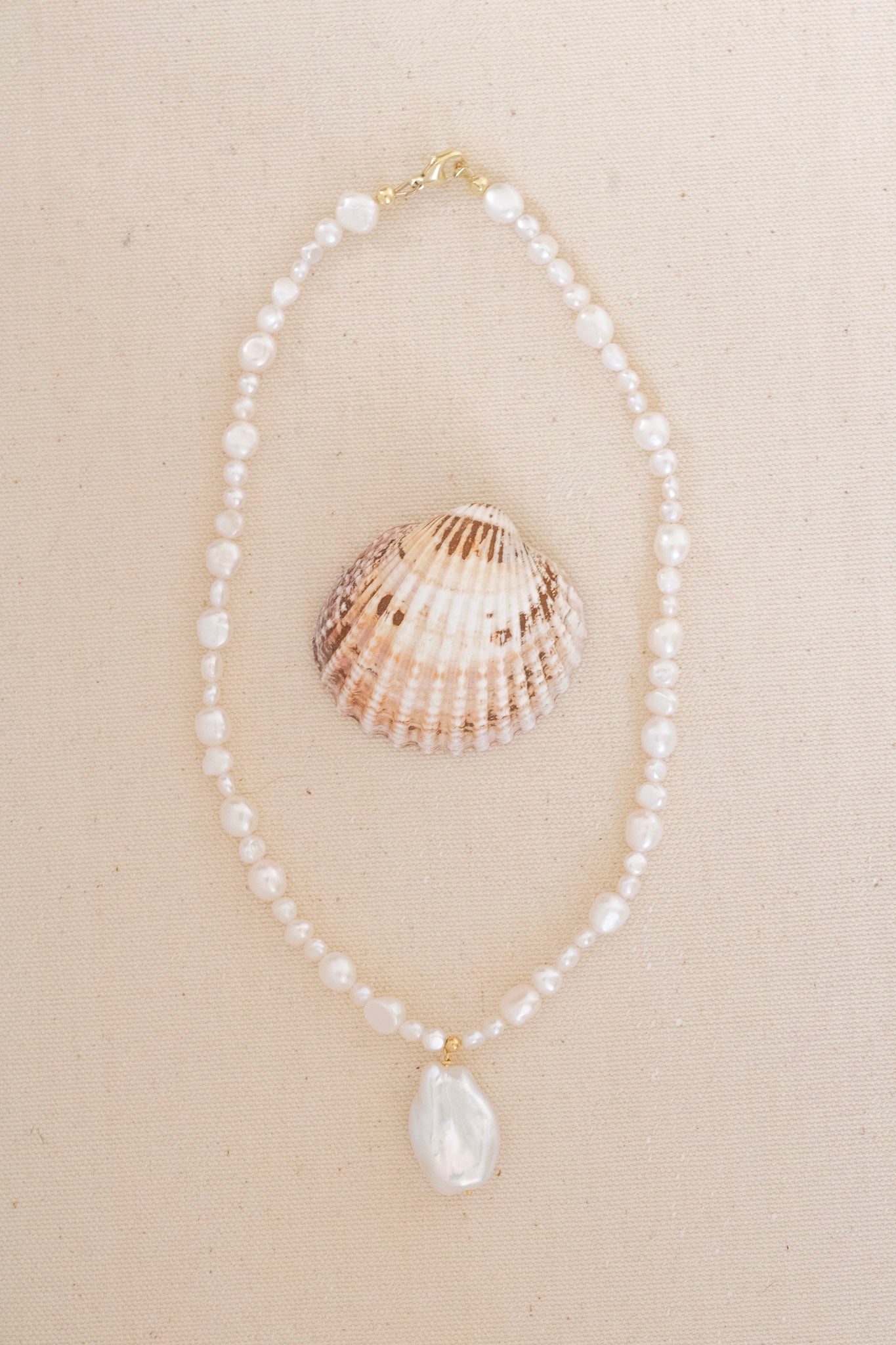 A delicate necklace made of small white pearls with a larger baroque pearl pendant, displayed on a beige textured background next to a seashell.