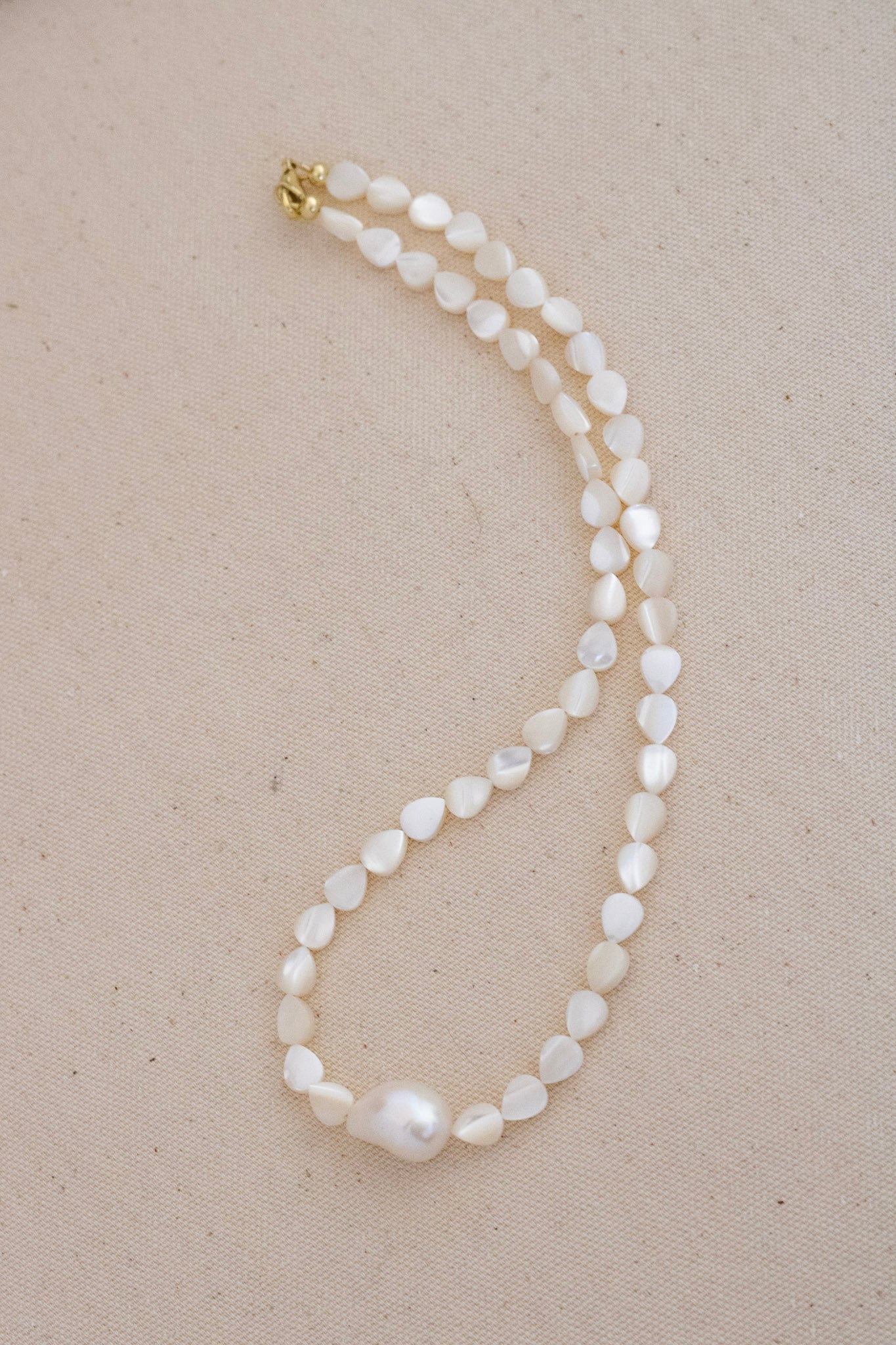 A single-strand baroque pearl necklace with a gold clasp, displayed on a textured beige fabric background.