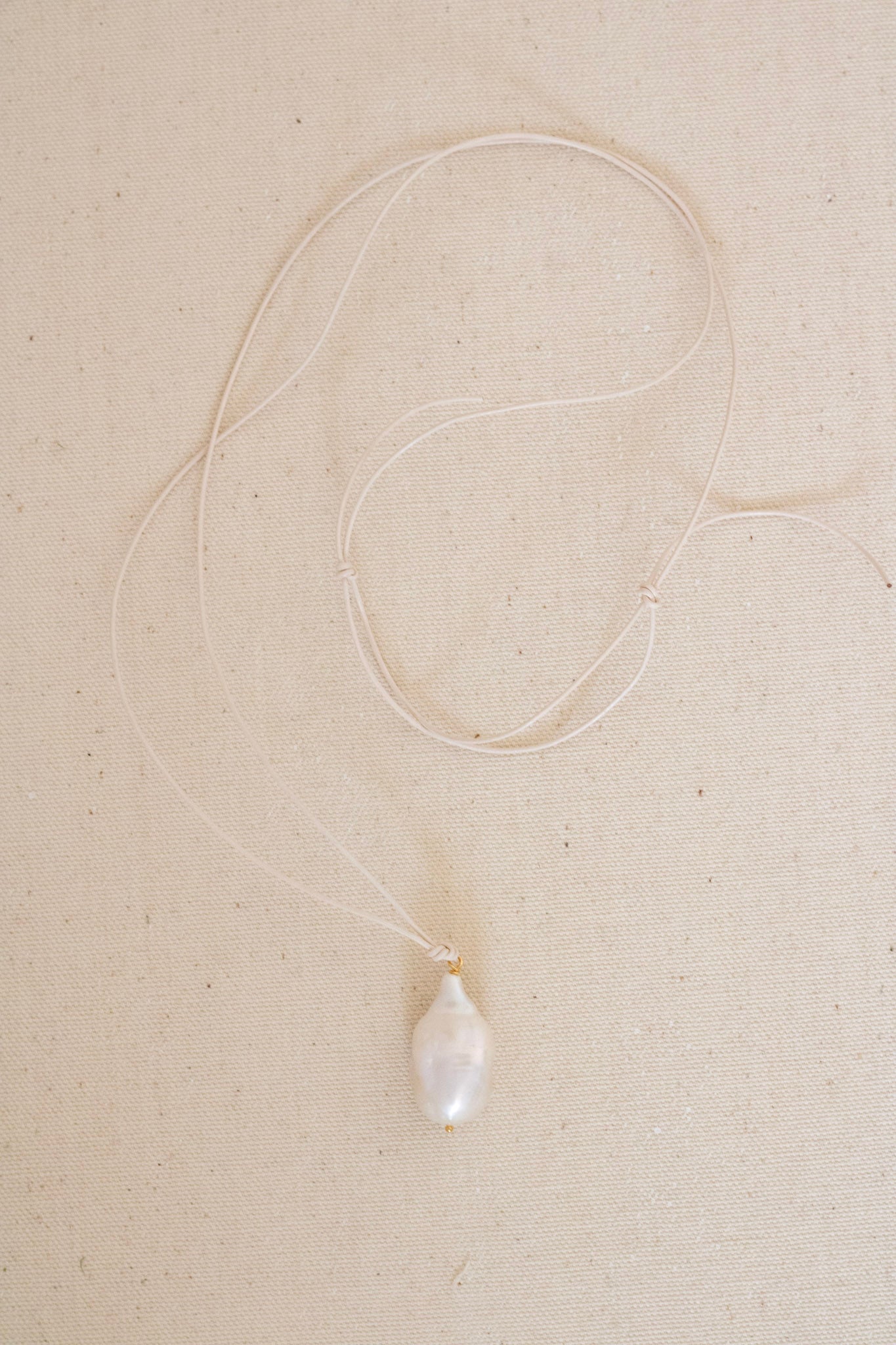 A delicate white cord necklace featuring a single irregular, baroque-style white pearl pendant, laid out on a textured beige fabric background.