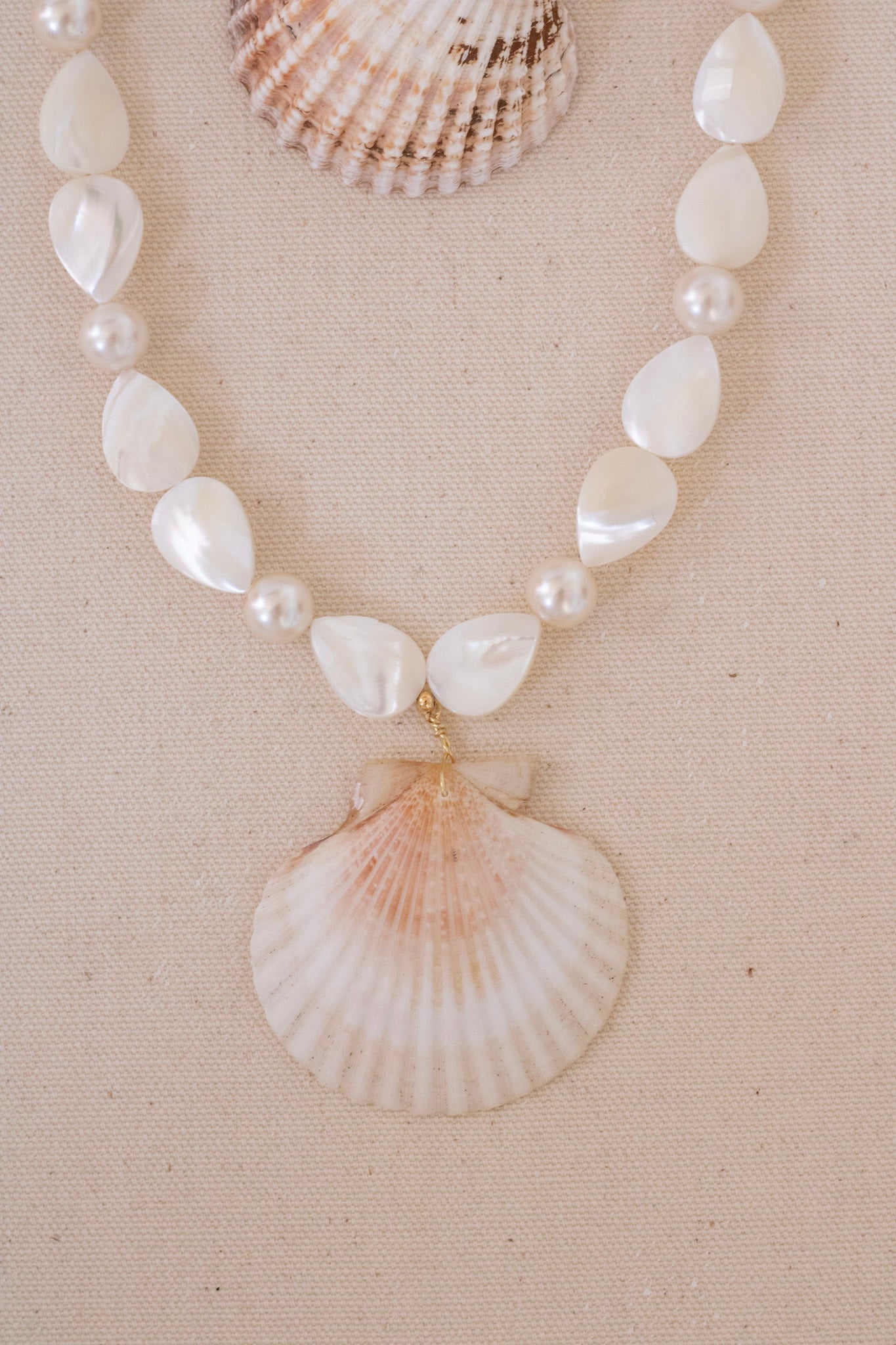 A shell-bead necklace displayed on a beige fabric background, featuring white teardrop-shaped shell beads, small pearl-like beads, and a large scallop shell pendant.