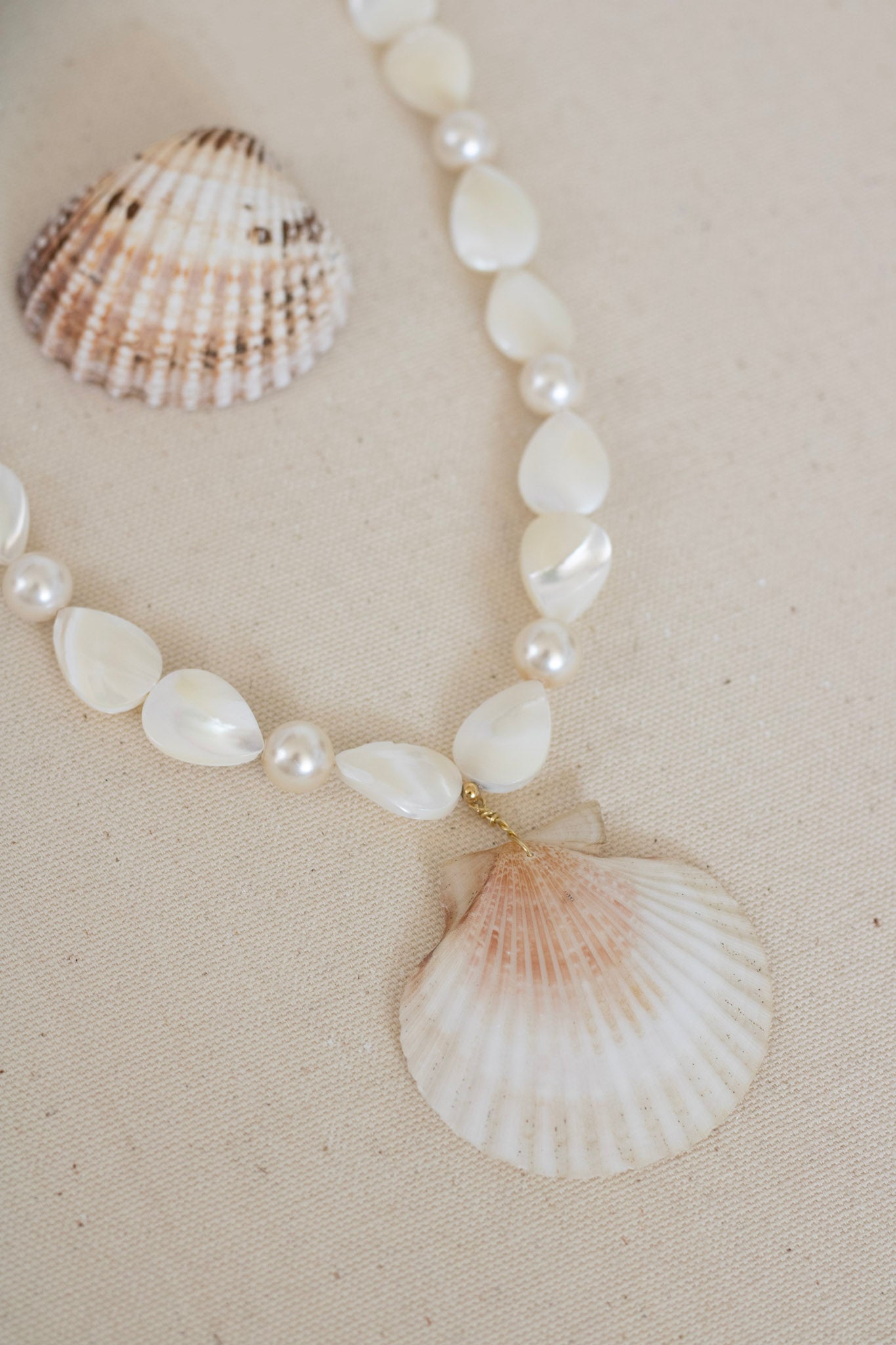 A beaded necklace featuring white mother-of-pearl teardrop beads, small white pearls, and a large scallop shell pendant, displayed on a beige textured fabric next to a seashell.