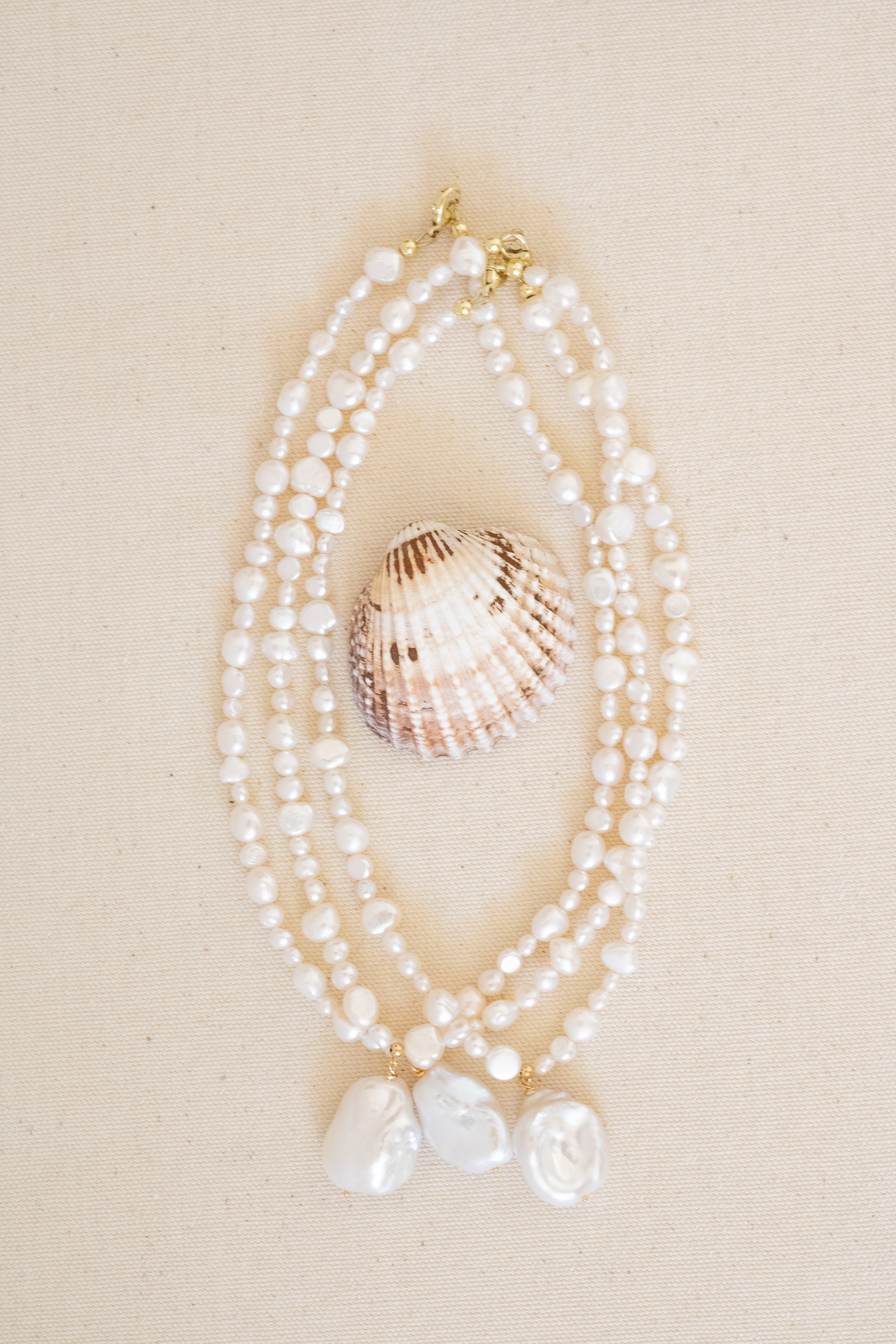 A multi-strand pearl necklace with gold-tone hardware and three larger baroque-style white pearls at the bottom, styled with a seashell on a beige fabric background.