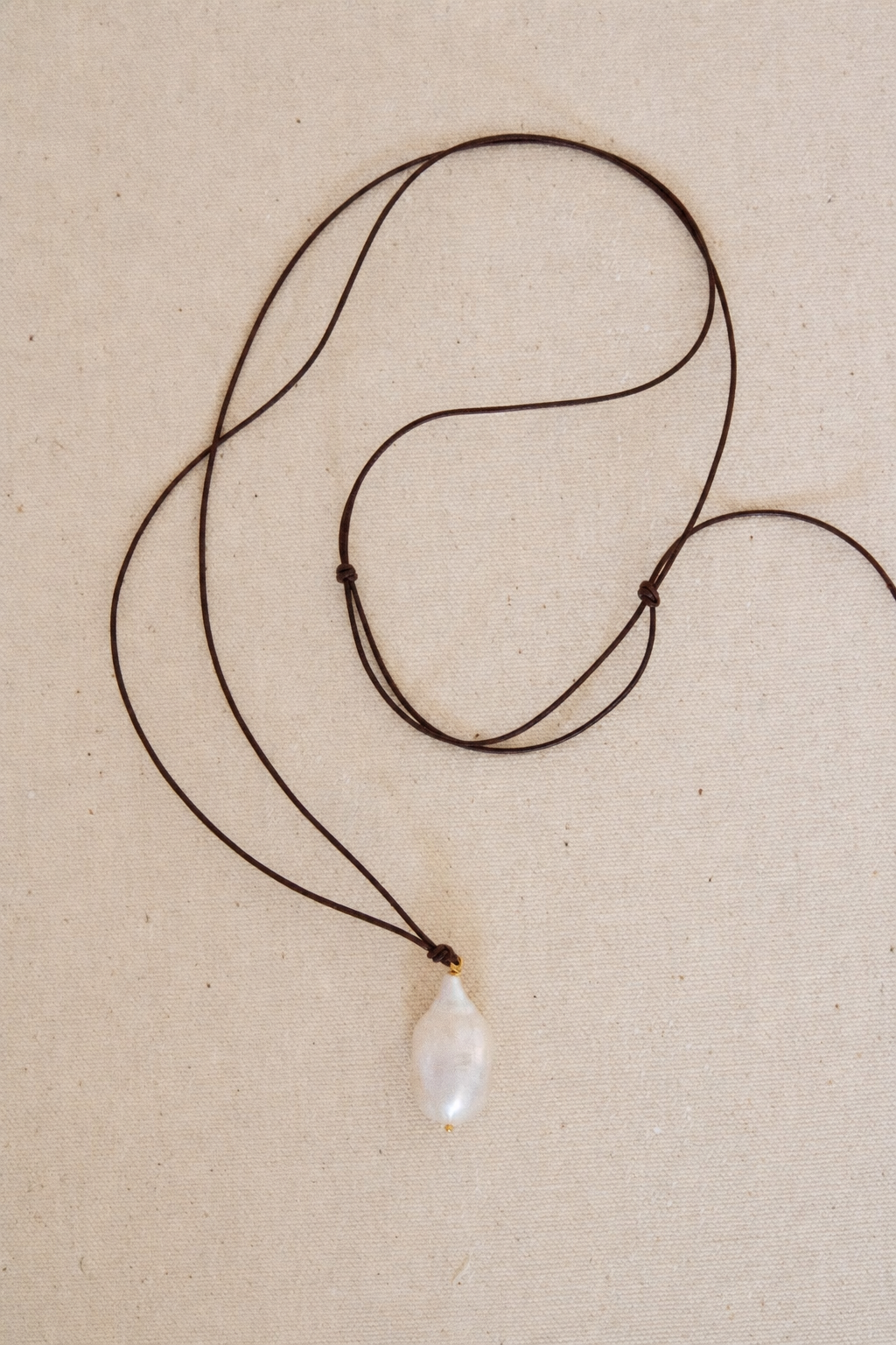 A dark brown cord necklace with a single irregular white pearl pendant, laid flat on a textured beige background.