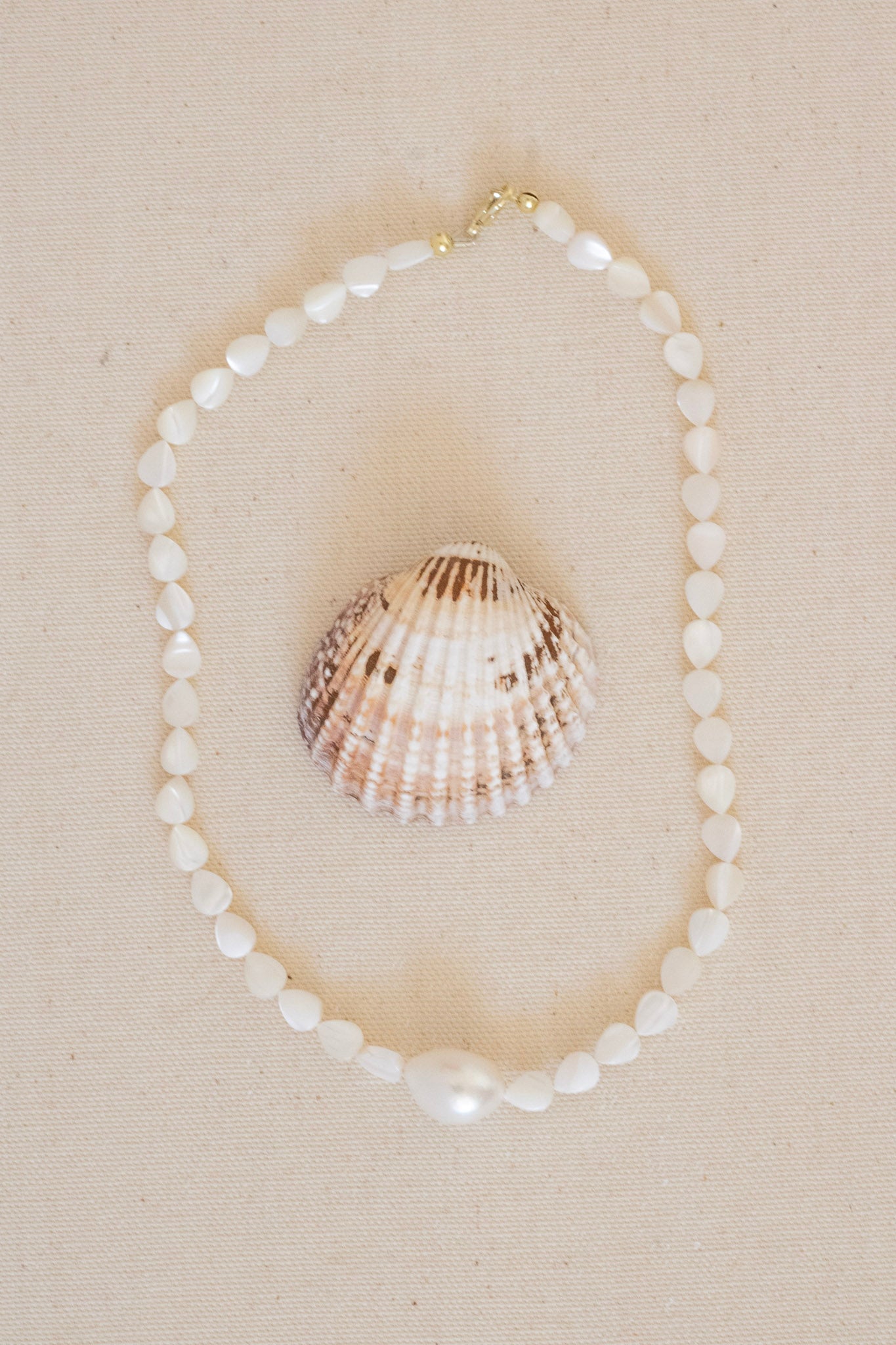 A white pearl and shell beaded necklace arranged in a circle on a beige textured fabric background, with a larger seashell placed in the center.