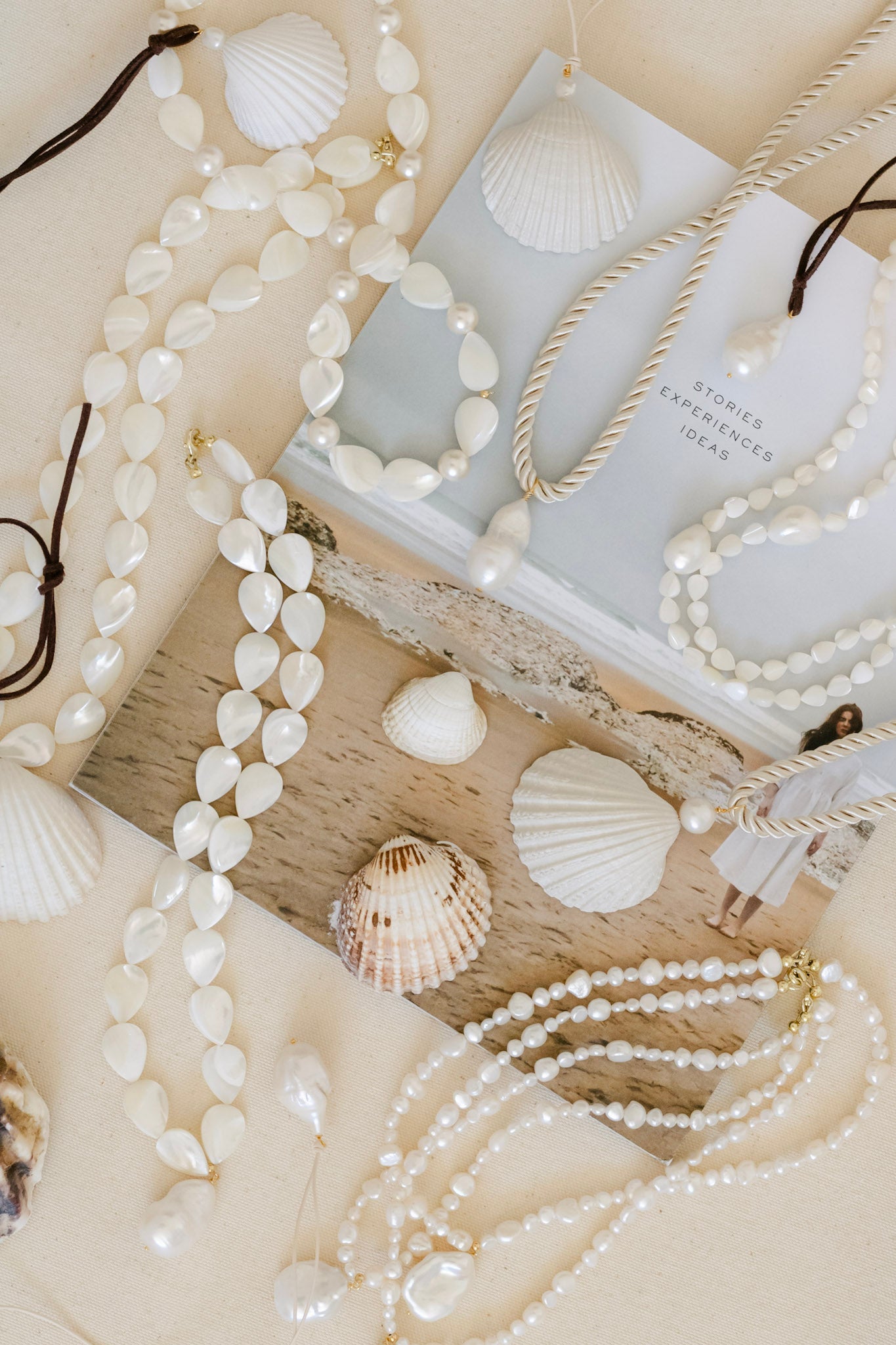 A styled flat-lay of multiple pearl and shell bead necklaces and bracelets arranged on a beige surface with seashells, a piece of driftwood, and a book.