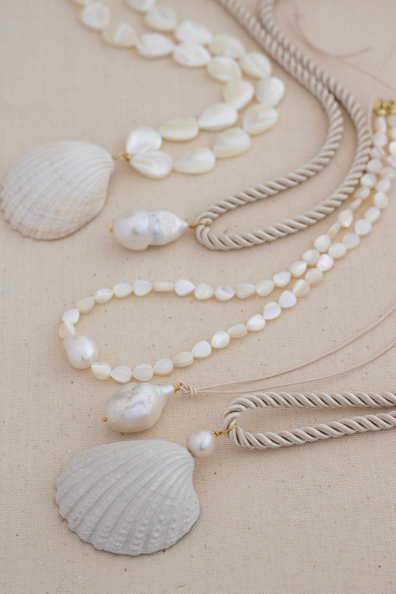 A set of bohemian-style layered necklaces featuring white seashells and baroque pearls on twisted beige cords, arranged on a neutral beige background.