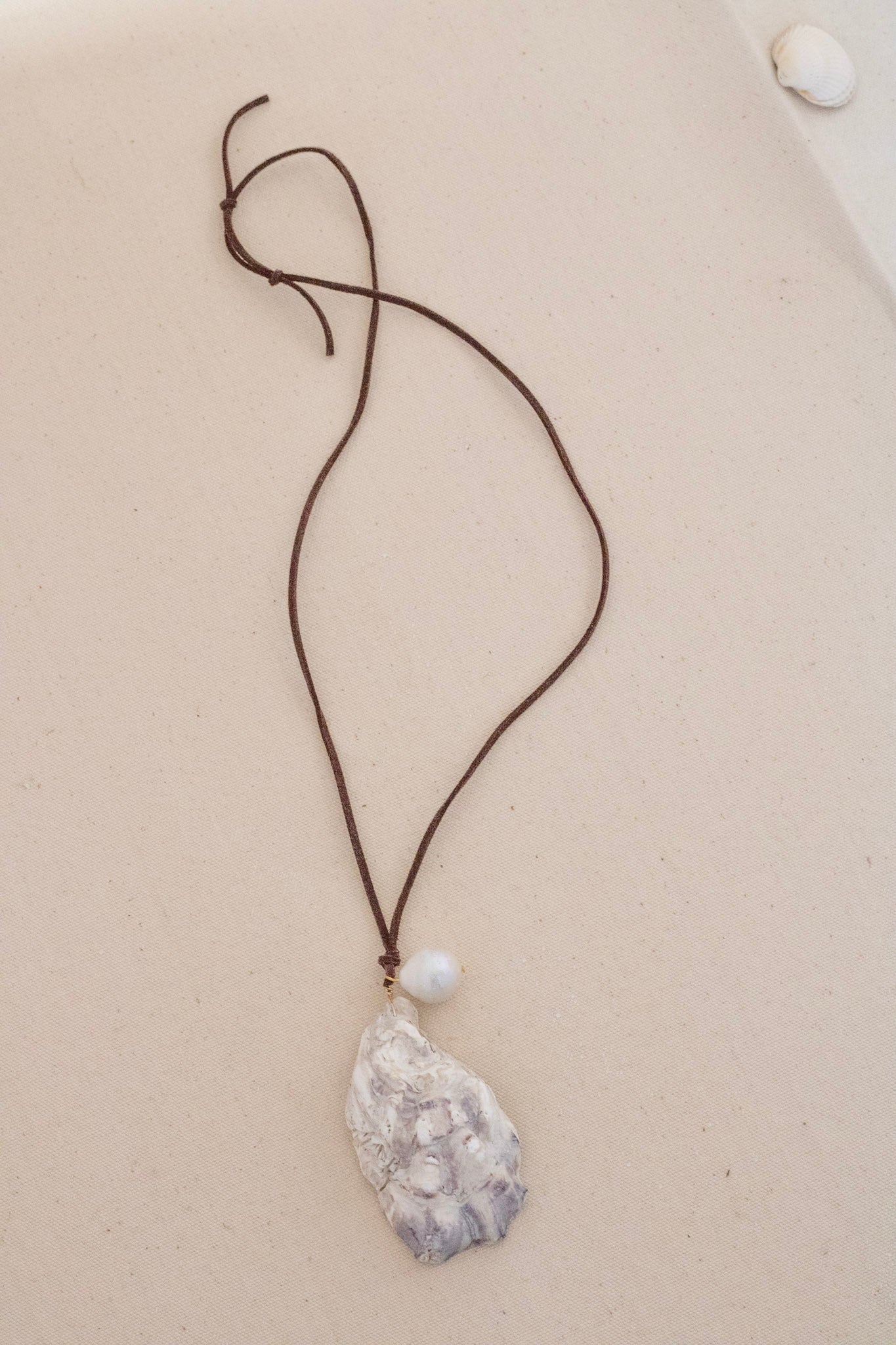 A necklace featuring a textured, organic-shaped white pendant with gray marbling, suspended from a dark brown cord with a small white pearl accent, displayed on a light beige surface.