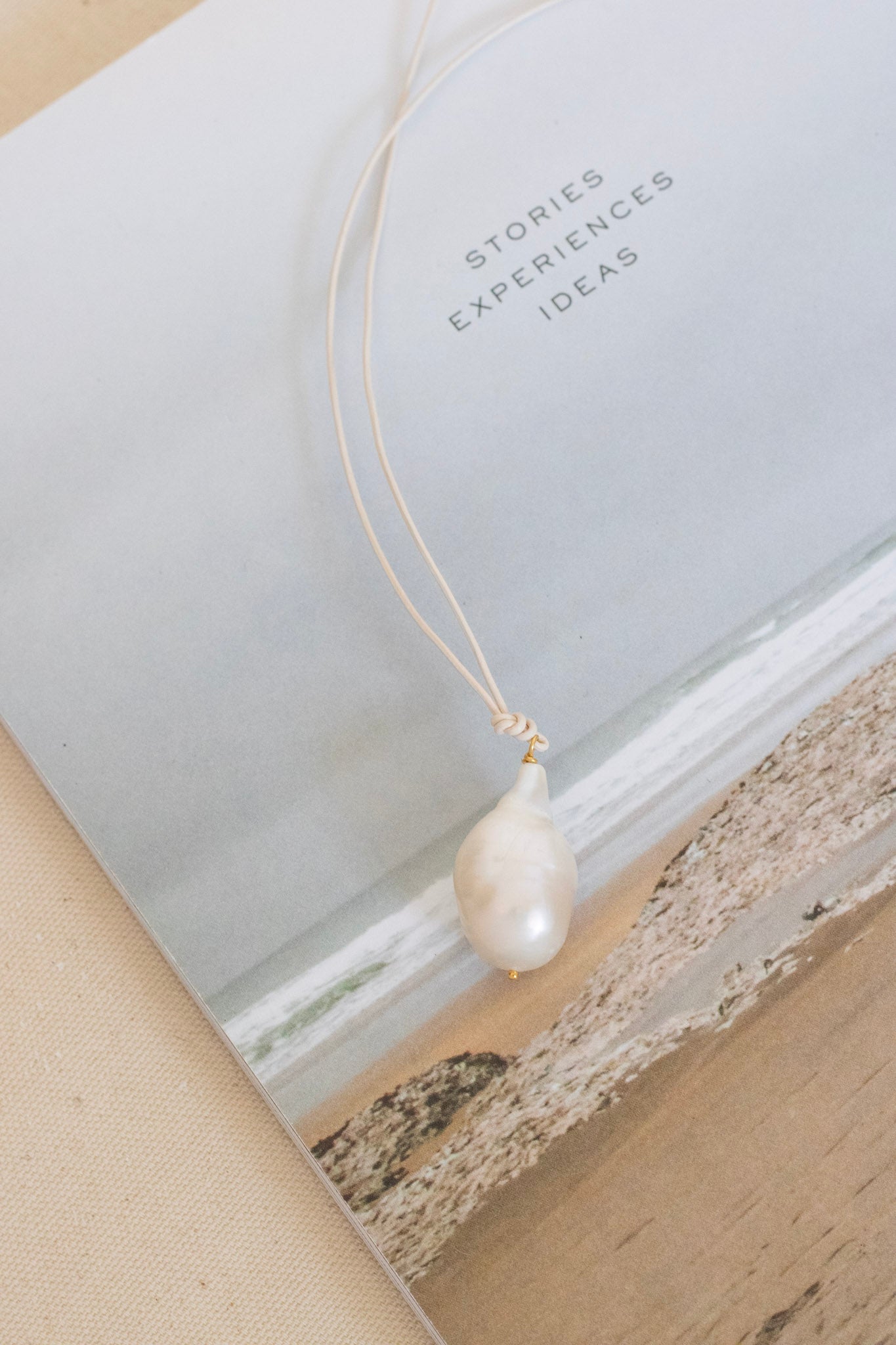 A delicate cream cord necklace with a single teardrop-shaped white freshwater pearl pendant, styled on top of a book with a beach illustration and the text 'STORIES EXPERIENCES IDEAS'.