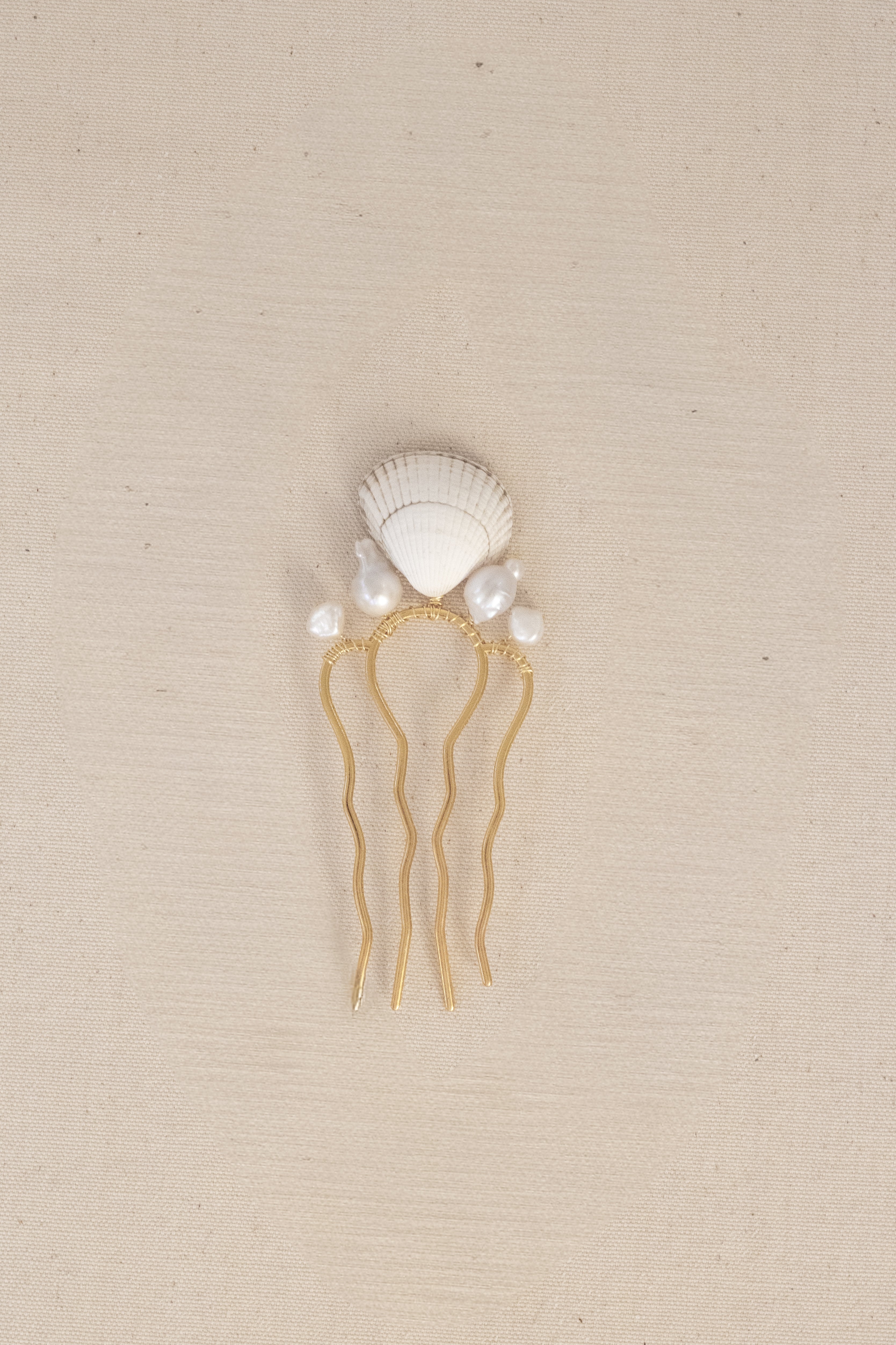 A gold hair comb featuring a white seashell on top, accented with small white pearls, set against a textured beige background.